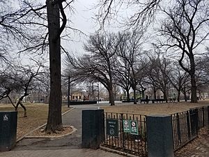 A green fence, about four feet high, opens on a path towards a basketball court, slightly up a hill. In the midground and background, atop lawns of brownish grass stand leafless trees against an overcast sky.