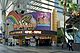 Ground-level view of the front entrance to the casino when it was called Fitzgeralds; a very prominent, colorful sign features a rainbow and a pot-of-gold. There are slot machines inside the building, and glass windows and part of a skyscraper are visible above the sign.