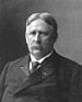 Head and shoulders of a white man with hair parted in the center and a mustache, wearing a dark suit and tie.