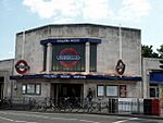 A light-grey-bricked building with a blue sign reading "COLLIERS WOOD STATION" in white letters all under a blue sky with white clouds