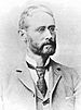 Head and shoulders of a white man with a full beard and wire-framed glasses, wearing a suit coat, vest, and tie.