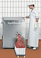 A man stands before a chest-high boxy machine that is pushing meat into an overflowing plastic bin on wheels.