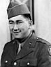 Head and shoulders of a smiling young man with dimples and round wire-framed glasses wearing a garrison cap and a military jacket over a shirt and tie.