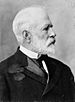 Profile of an older, balding white man with a short-cropped white beard, wearing a dark suit jacket over a white shirt and dark tie.