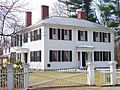 A white two story house, whose main section is square, although there is a wing extending off the back. There is a white fence with a gate across a gravel drive.